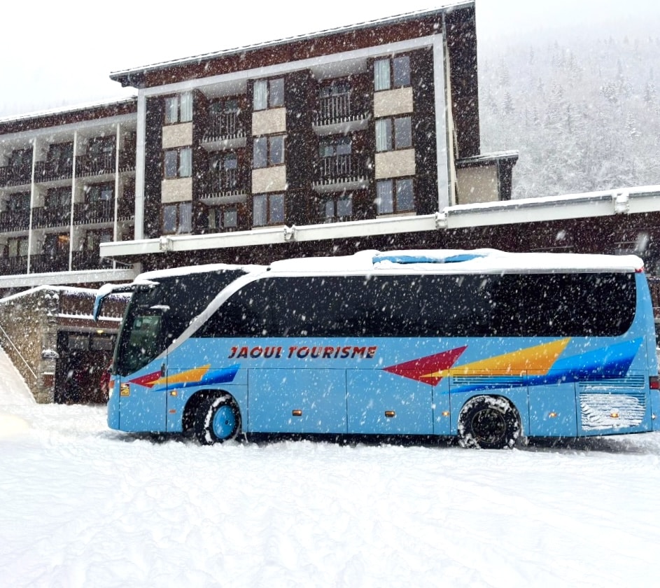 Autocar bleu Jaoul Tourisme devant un hôtel enneigé lors d'une chute de neige en montagne.