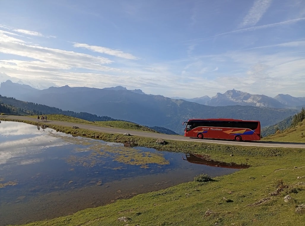 Bus rouge Inoui Tourisme au bord d'un lac alpin avec vue panoramique sur les montagnes.