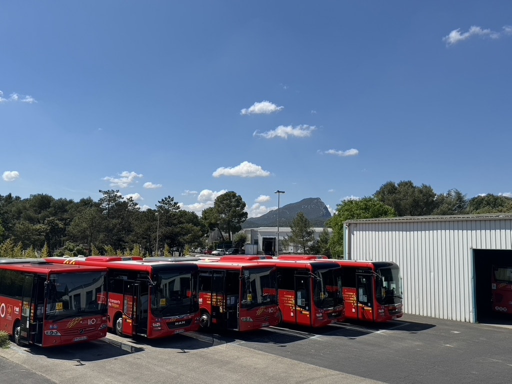 Bus rouges liO et MAN stationnés devant un entrepôt blanc avec montagnes en arrière-plan.